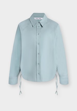 Ανοιχτό μπλε μακρυμάνικο πουκάμισο με κουμπιά, μυτερό γιακά και πλαϊνά κορδόνια, απλωμένο επίπεδα σε λευκό φόντο.