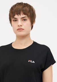 Melna īsām piedurknēm T-kotetes, izgatavotas no kokvilnas, ar apaļu kakla izgriezumu un FILA logo, kas izšūts balti un sarkani uz kreisā krūšu sāna.