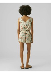 Nainen, jolla on kiharat hiukset, pukeutuneena olkaimeton kukkakuosiseksi toppiin ja samankaltaisiin shortsseihin, seisoo paljain jaloin tasaisella taustalla, kasvot poispäin.