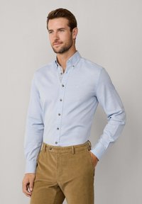 Camisa de botones azul claro con un cuello interior a cuadros, botones marrones y mangas largas, combinada con pantalones de corte de pana color beige.