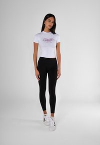 Hvit avkortet t-skjorte med teksten "Lucky Girl" og sommerfugl grafikk, kombinert med svarte leggings og hvite sneakers med svarte striper.