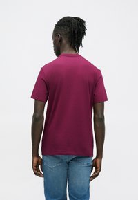 Bordowa koszulka z krótkim rękawem wykonana z miękkiego materiału. Posiada klasyczny okrągły dekolt. Noszona z niebieskimi dżinsami. Widok od tyłu.