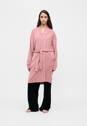 Femme debout pieds nus portant une robe de chambre rose nouée à la taille et un pantalon noir ample sur un fond blanc uni.