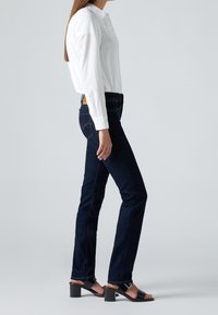 Femme en chemise blanche à manches longues, jean bleu foncé et sandales noires à talons, debout de profil sur un fond gris uni.