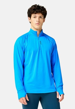 Giovane uomo che indossa una maglietta sportiva blu brillante a maniche lunghe con zip parziale e pantaloni neri su uno sfondo chiaro e uniforme.