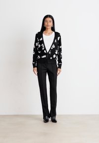 Svart kofta med vitt abstrakt blommönster, V-ringning, frontknappar, kombinerad med svarta skräddarsydda byxor, bärs över en vit topp.