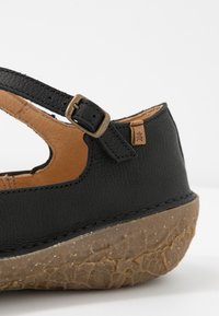 Svart lädersandal med en texturerad yta, justerbar rem med ett metallspänne och en naturlig, grov sula.