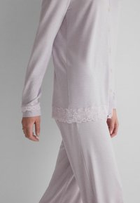 Person trägt ein helllila Freizeitkleid-Set mit einem knöpfbaren Oberteil, das Spitzenbesatz an Manschetten und Saum hat, und passende Hosen.