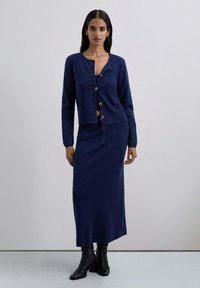 Cardigán de punto azul marino con botones dorados, combinado con una falda larga azul marino. Textura suave, diseño ajustado y botas negras hasta la rodilla.