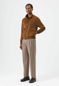 Brown suede jacket with a high collar, front buttons, and a pocket; paired with light beige trousers and dark brown shoes.