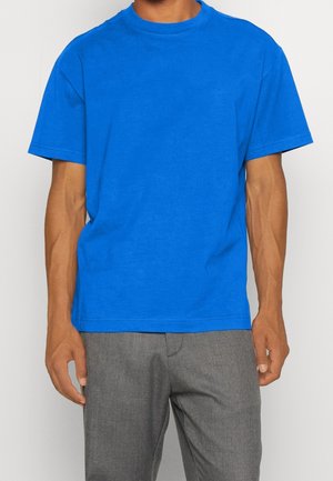 Niebieska bawełniana koszulka z okrągłym dekoltem, krótkimi rękawami i luźnym krojem, zestawiona z szarymi teksturowanymi spodniami. Brak widocznych logo lub wzorów.