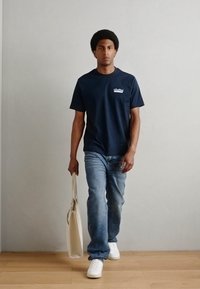 Marine T-skjorte med logo på venstre bryst, blå jeans og hvite joggesko. Holder en beige tote bag, står på tregulv mot en lys vegg.
