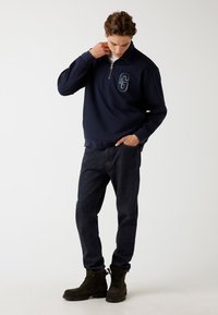 Marineblauwe fleece pullover met een ritskraag en een verhoogd "G"-logo op de borst, gecombineerd met donkere spijkerbroeken en bruine veterschoenen.