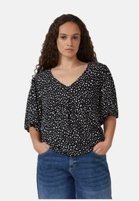 Femme aux cheveux bouclés portant un blouse noire à d�écolleté en V avec des pois blancs et des jeans bleus, les mains dans les poches, contre un fond clair.