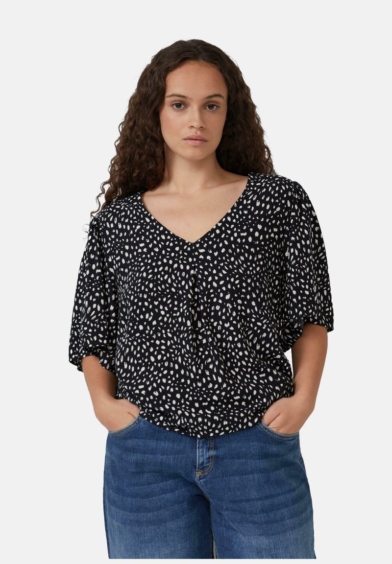 Femme aux cheveux bouclés portant un blouse noire à décolleté en V avec des pois blancs et des jeans bleus, les mains dans les poches, contre un fond clair.