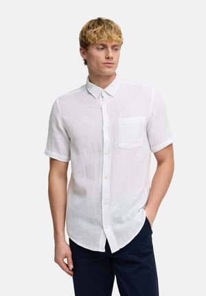 Vaaleahiuksinen mies, joka pitää valkoista lyhythihaista nappipaitaa, jossa on rintatasku, ja tummansiniset housut, seisoo yhdellä kädellä taskussa.