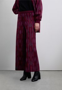 Pantaloni a gamba larga in maglia color borgogna con un motivo geometrico rosso e grigio. Abbinati a stivaletti neri, con una trama intricata visibile.