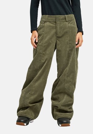 Personne portant un pantalon large en velours côtelé vert olive avec des poches, des chaussures noires et un haut noir à manches longues sur un fond uni.