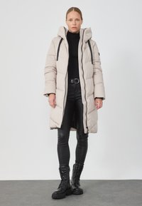 Puffjakke i lys beige, med hette, glidelås og svarte stropper. Sammen med svarte skinnleggings og svarte snørestøvler.