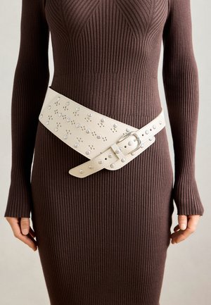 Witte leren riem met studs, met een brede ontwerp en een rechthoekige gesp, gedragen over een aansluitende bruine geribbelde jurk.