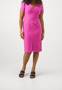 Femme portant une robe rose ajustée jusqu'aux genoux avec des manches courtes et des sandales noires à bouts ouverts, debout devant un fond blanc uni.