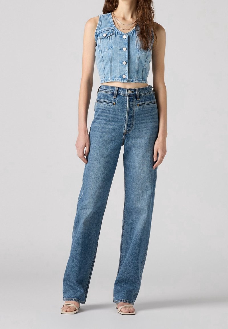 Femme portant un gilet en jean court sans manches avec des boutons sur le devant et un jean bleu taille haute droit, debout devant un fond uni.