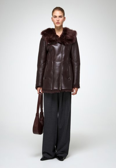 Giacca in pelle marrone con colletto in pelliccia, dotata di chiusura con zip, tasche laterali e un design elegante, abbinata a pantaloni grigi a gamba larga e una borsa.