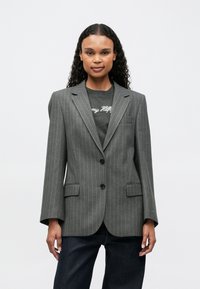 Blazer gris a rayas con un corte entallado, solapas con muescas, dos bolsillos frontales y dos botones. Lleva puesto un camiseta gráfica oscura.