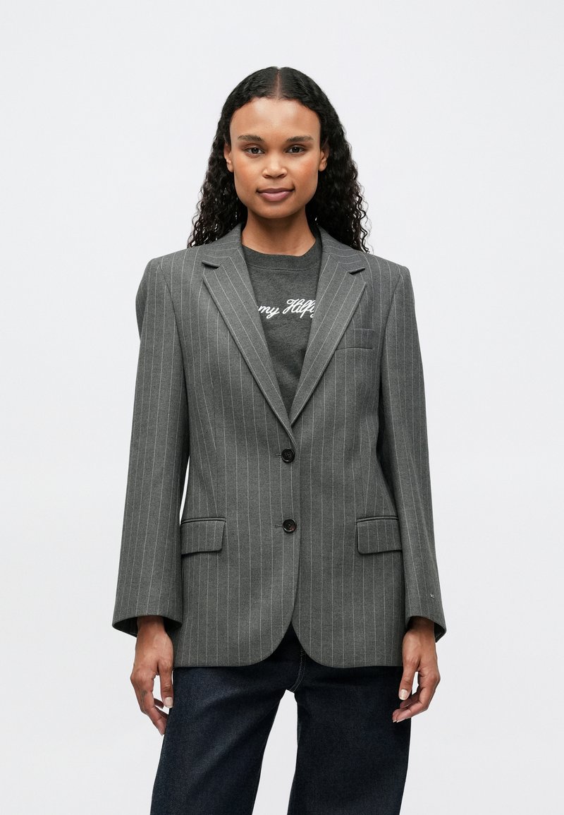 Blazer gris a rayas con un corte entallado, solapas con muescas, dos bolsillos frontales y dos botones. Lleva puesto un camiseta gráfica oscura.