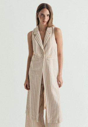 Mujer con chaleco largo sin mangas a rayas verticales beige y blancas con botones, de pie frente a un fondo claro y liso.