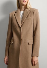 Taupe wollen jas met een gestructureerd ontwerp, geknipte revers, twee voorzakken en een gedetailleerde borstzak. Zachte textuur, getailleerde pasvorm.