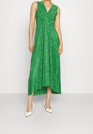 Mannequin portant une robe verte plissée sans manches, à col en V et à l’ourlet asymétrique, avec des escarpins blancs à bout pointu.
