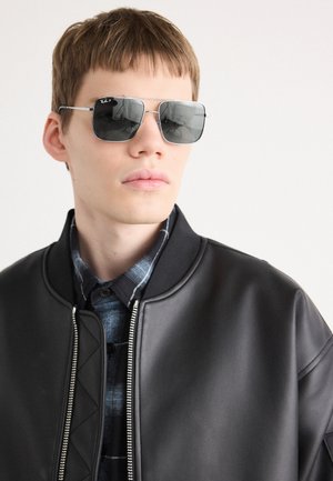 Sonnenbrille mit dunklen Gläsern, silbernem Metallrahmen, rechteckiger Form. Das Modell trägt eine schwarze Lederbomberjacke über einem blauen Karoshemd.