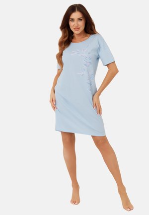 Kobieta stojąca boso, ubrana w jasnoniebieską sukienkę do kolan z krótkimi rękawami i wzorem różowo-niebieskich liści na przodzie.