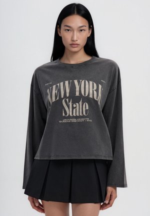 Giovane donna con capelli lunghi e neri che indossa una maglietta a maniche lunghe grigio scuro con testo "New York State" e una gonna plissettata nera.