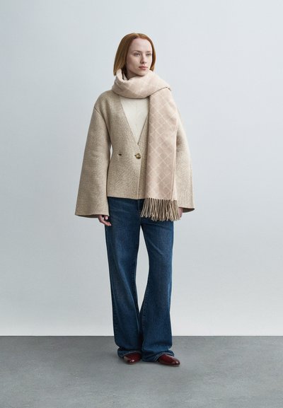 Γυναίκα που φοράει μπεζ ζακέτα, φουλάρι με το ίδιο σχέδιο, κρεμ μπλούζα, τζιν παντελόνι με φαρδιά πόδια και καφέ παπούτσια πάνω σε απλό γκρι φόντο.