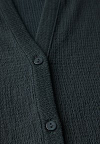 Mörkgrön texturerad cardigan med V-ringning och en enda svart knappstängning. Tyget är vågigt med en mjuk finish.