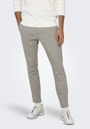 Mann trägt hellgraue Slim-Fit-Hosen und weiße Sneaker mit Socken, Hände teilweise in den Taschen vor einfachem Hintergrund.