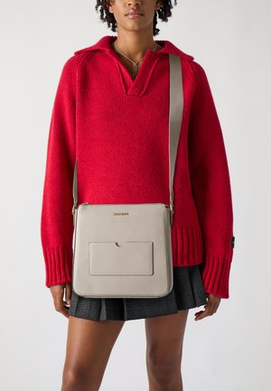 Borsa a tracolla grigia in pelle con tasca frontale, forma rettangolare e tracolla regolabile, abbinata a un maglione rosso lavorato a maglia e una gonna a quadri.