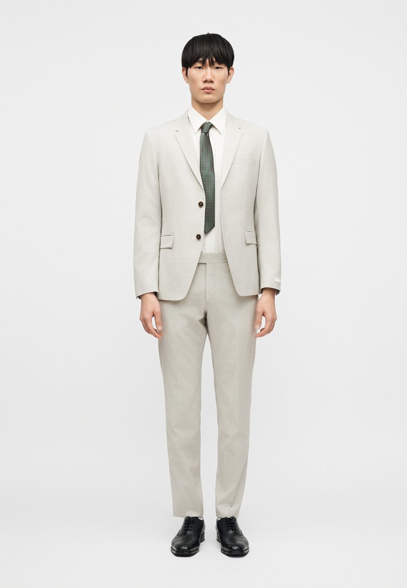 Ανοιχτό γκρι κοστούμι που αποτελείται από ένα εφαρμοστό σακάκι και κωνικά παντελόνια, συνδυασμένο με λευκό πουκάμισο και σκουρό πράσινη γραβάτα, συμπληρωμένο με μαύρα παπούτσια.