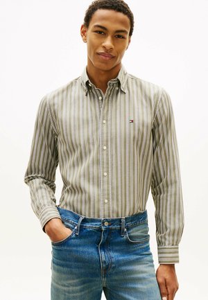 Junger Mann, der ein grün-weiß gestreiftes Hemd mit Knöpfen trägt, das in blaue Jeans gesteckt ist, steht mit einer Hand in der Tasche vor einem schlichten Hintergrund.