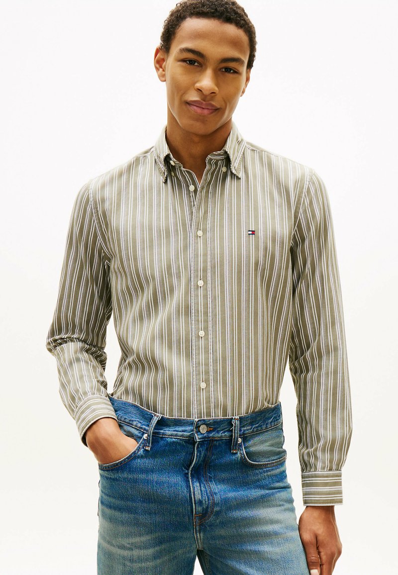 Jeune homme portant une chemise à rayures vertes et blanches rentrée dans un jean bleu, debout avec une main dans la poche sur un fond uni.