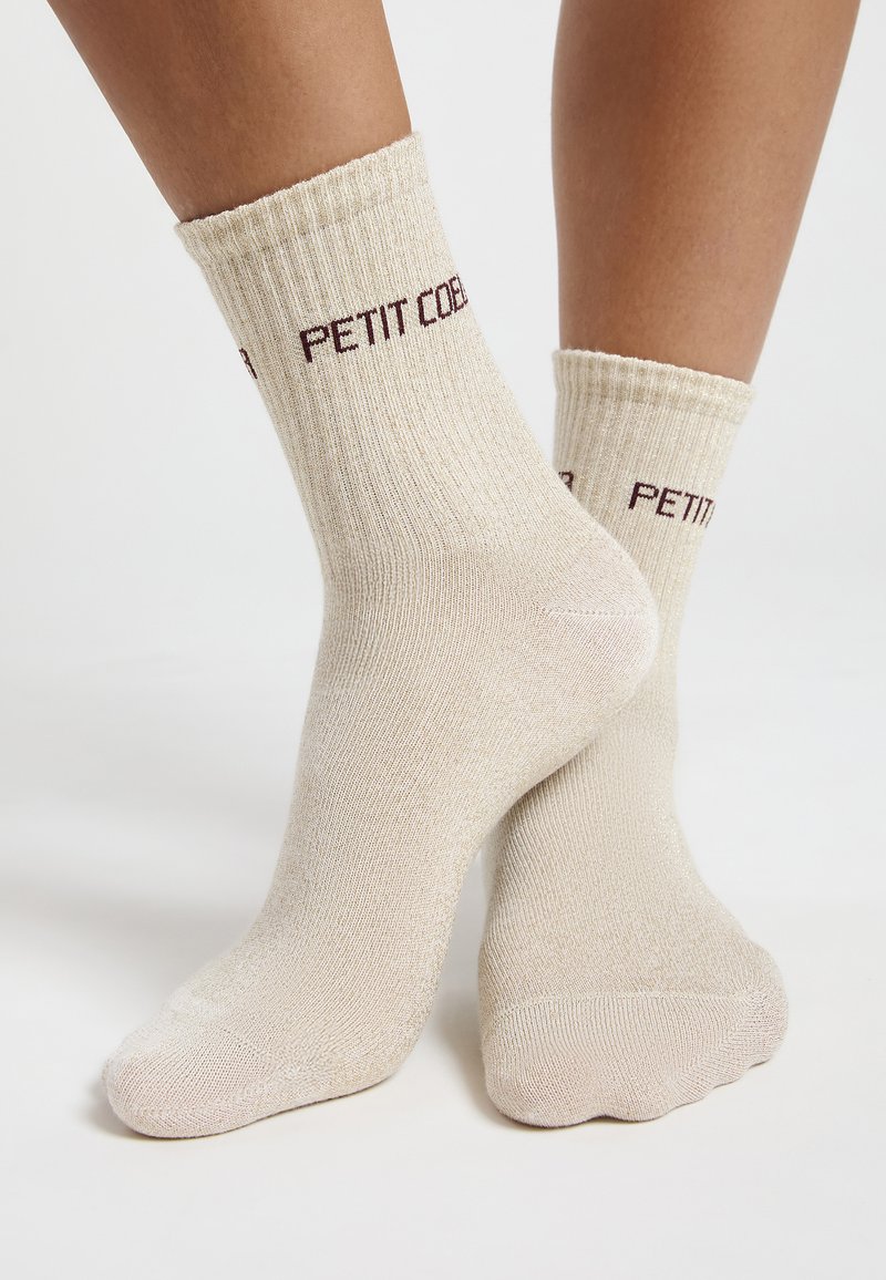 Beige gerippte Socken mit einem umgeschlagenen Saum, versehen mit burgunderrotem Text "PETIT COBER." Das Material wirkt weich mit einer dezenten Textur.