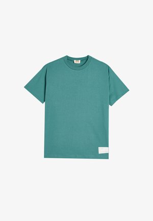 Camiseta de algodón color teal con mangas cortas, cuello redondo y una etiqueta rectangular en la parte inferior que muestra el texto "el océano".
