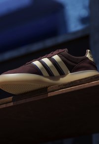 Vue en contre-plongée d'une basket marron avec des rayures crème et une semelle reposant sur une surface en bois devant un fond bleu foncé.