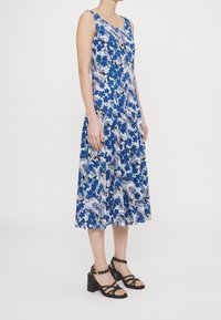 Robe fleurie avec motifs bleus et blancs, design sans manches, silhouette en A, associée à des sandales à talons carrés noires.