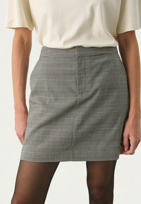 Siva karirana suknja ima uski kroj s visokim strukom. Materijal izgleda teksturirano s suptilnim linijama, uparen s prozirnim crnim pantyhose.