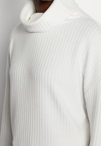 Weißer, gerippter Rollkragenpullover aus dickem, strukturiertem Material. Verfügt über einen breiten, umgeschlagenen Kragen und eine locker sitzende Passform.