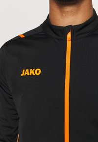 Svart idrottjacka med orange dragkedja och detaljer. "JAKO"-logotyp i orange på vänster bröst. Slät textur, lättviktig design.