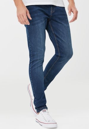 Mężczyzna noszący dopasowane niebieskie jeansy i białe niskie sneakersy, stojący z jedną lekko ugiętą nogą na jednolitym tle.